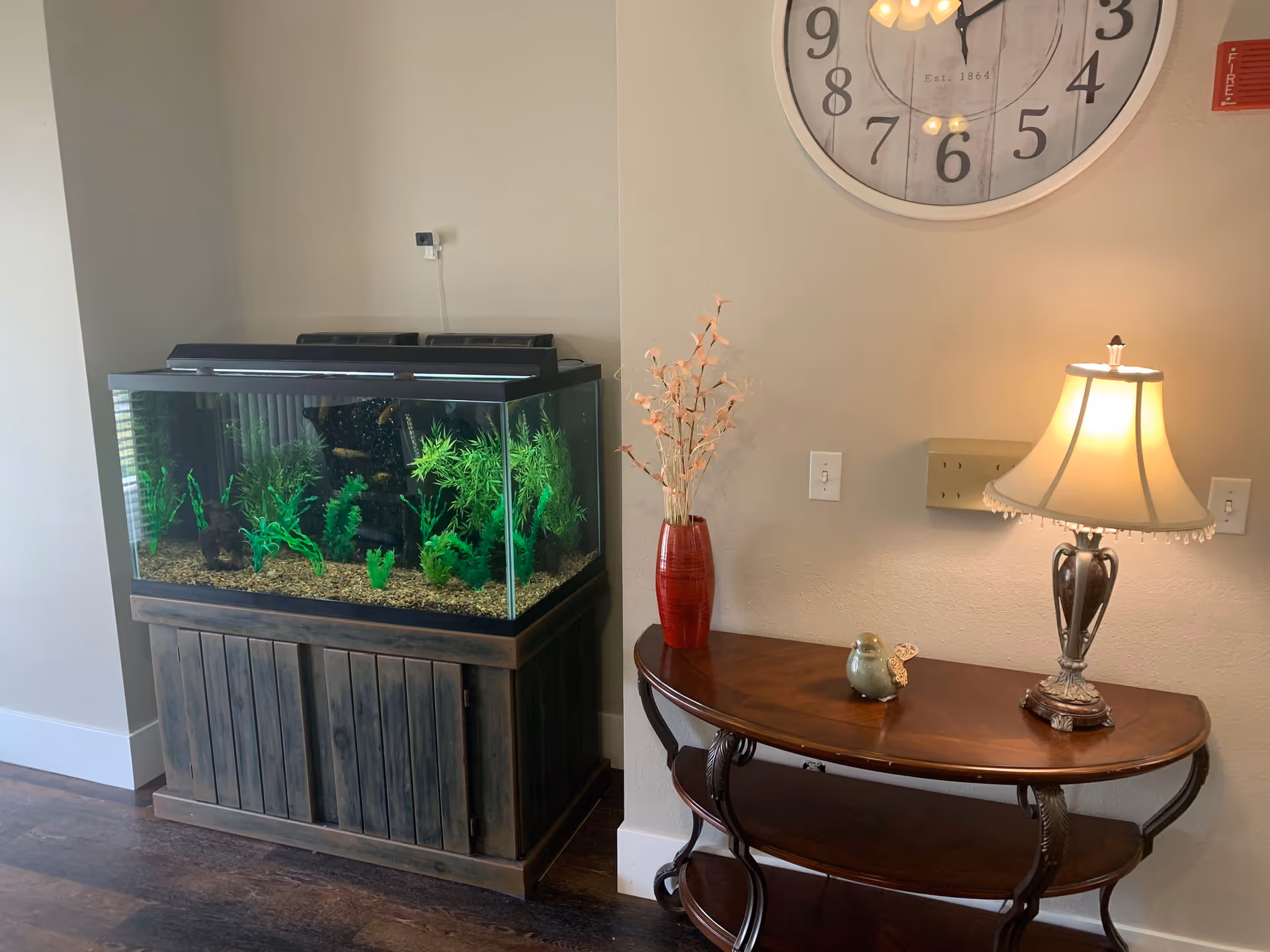 A corner of a room featuring a large fish tank with green aquatic plants on a wooden stand, next to a wooden console table with a red vase holding dried flowers, a small decorative bird, and a lit table lamp. Above the table is a large round wall clock and a fire alarm on the wall.