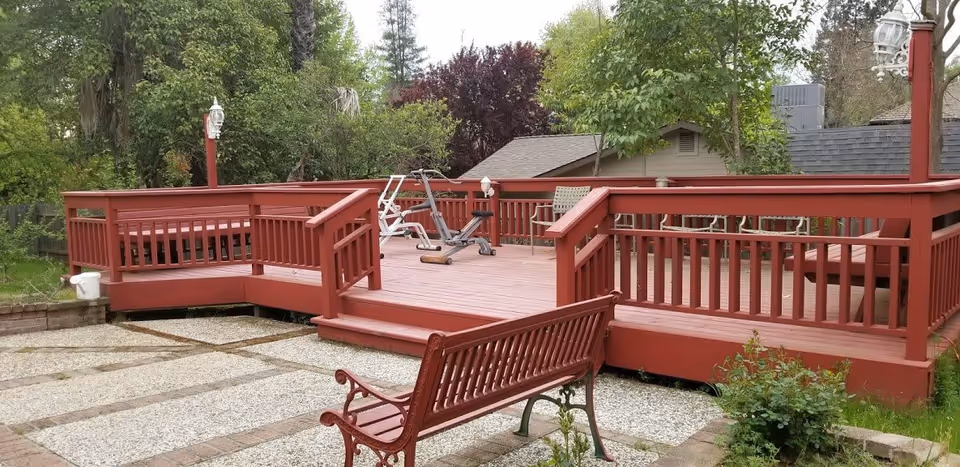 Outdoor wooden deck painted red with railings, a bench in the foreground, exercise equipment on the deck, surrounded by trees and greenery.