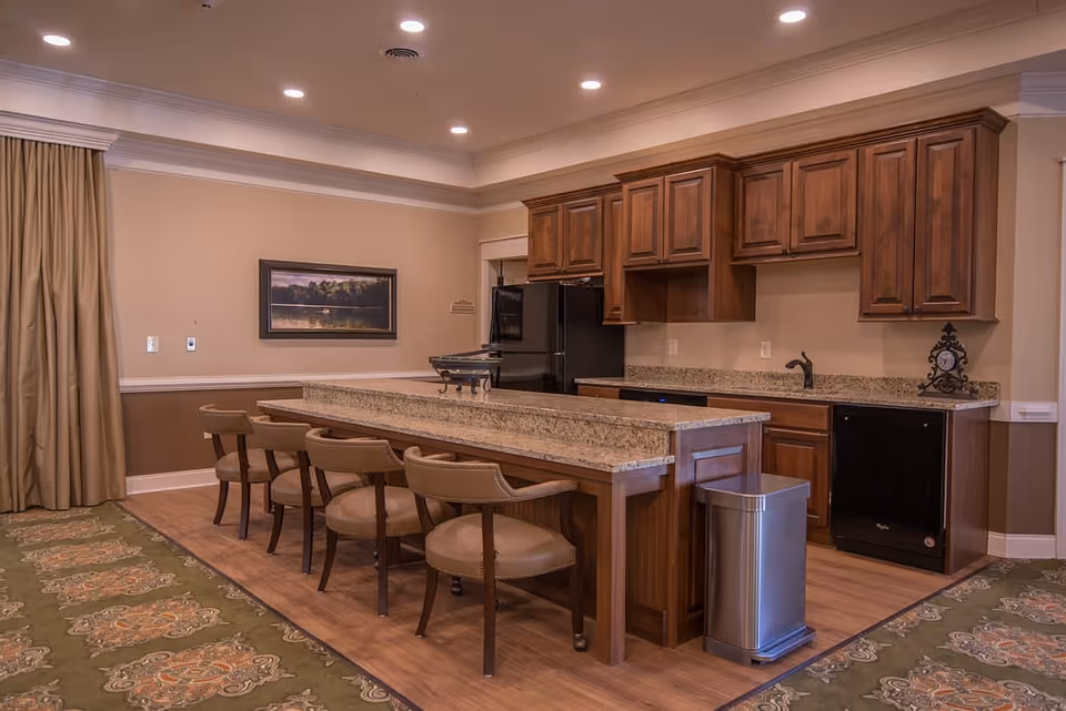 Community kitchen area with a long granite island, wooden cabinets, barstools and appliances.