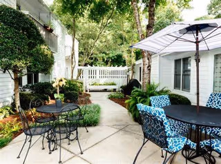Outdoor patio area with black metal tables and chairs, some with blue patterned cushions, and a large white umbrella. The patio is surrounded by greenery including bushes, trees, and plants, with a white fence and white building walls in the background.