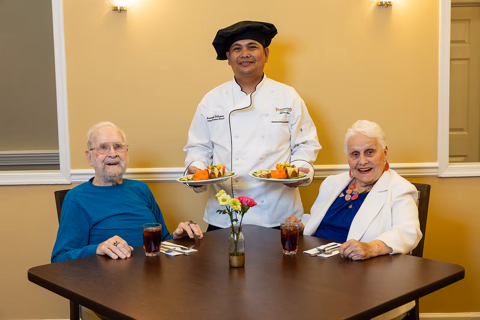A chef wearing a white uniform and black chef hat stands behind a dining table holding two plates of food. Seated at the table are an elderly man in a blue shirt and an elderly woman in a white jacket and blue top, both smiling. The table has two glasses of dark beverage, utensils, and a small vase with flowers. The background features a beige wall with white trim and two wall sconces.