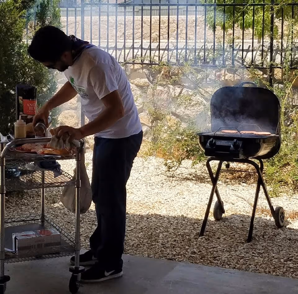A man wearing a white Good Life Assisted Living and Memory Care El Paso t-shirt is preparing food on a metal cart next to a smoking charcoal grill with burgers cooking outdoors in a fenced area with gravel ground and some greenery.