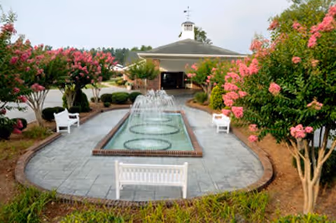 Outdoor courtyard area with a rectangular water fountain featuring multiple water jets, surrounded by paved walkways and white benches. Pink flowering trees and green shrubs line the sides, with a building visible in the background under a cloudy sky.