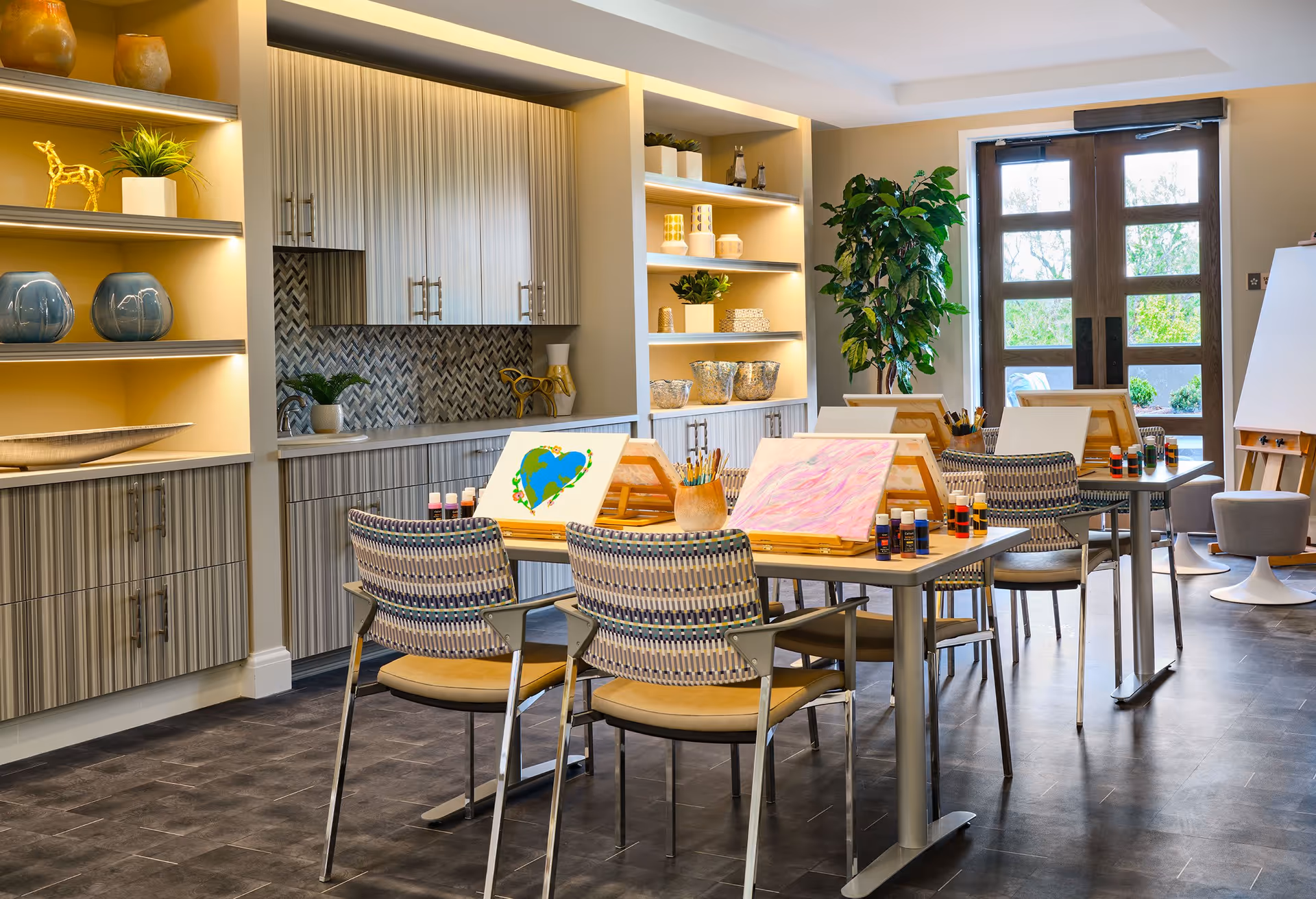 A bright and inviting art activity room with tables and chairs arranged for painting. Each table has easels with canvases, paint bottles, and brushes. The room features built-in shelves with decorative items, a large plant near glass doors letting in natural light, and cabinetry with a modern backsplash.