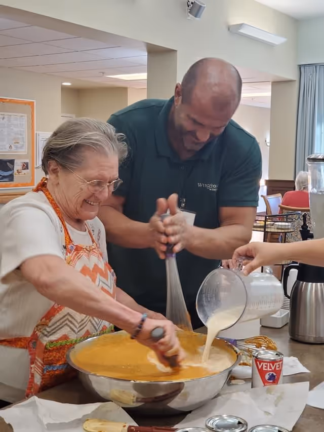 An elderly woman and a man are smiling and mixing ingredients in a large metal bowl in a kitchen or dining area. The woman is stirring with a spatula while the man is whisking. Another person is pouring a liquid from a measuring cup into the bowl. The man is wearing a green shirt with the WindSong at Southridge logo. The setting appears to be a communal or senior living facility kitchen or activity area.