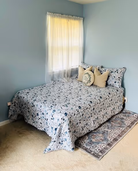 A cozy bedroom with light blue walls featuring a bed covered with a floral patterned bedspread in shades of blue and white. The bed has several decorative pillows, including a round one with a floral design. A sheer white curtain covers the window behind the bed, and a patterned rug is partially visible under the bed on a beige carpeted floor.