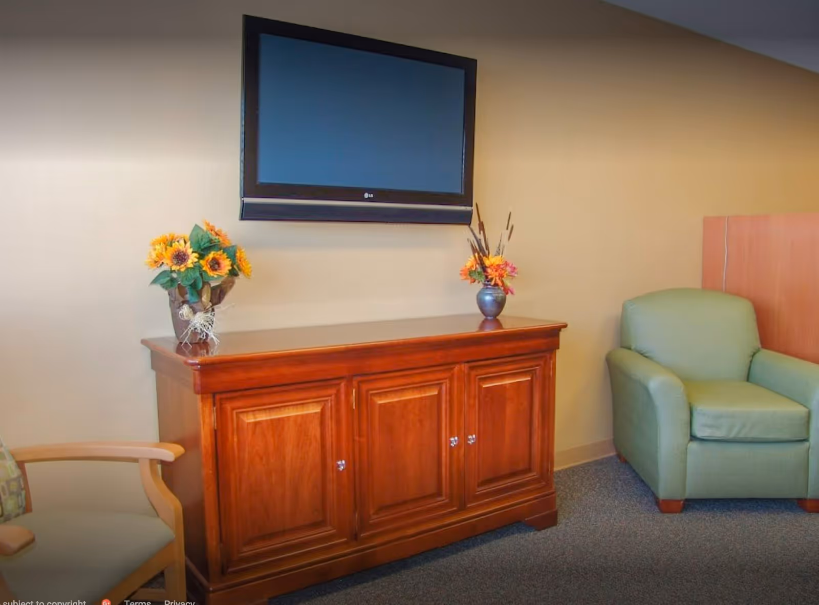 Wall-mounted TV above a wooden cabinet with floral arrangements and upholstered chairs in a sitting area.