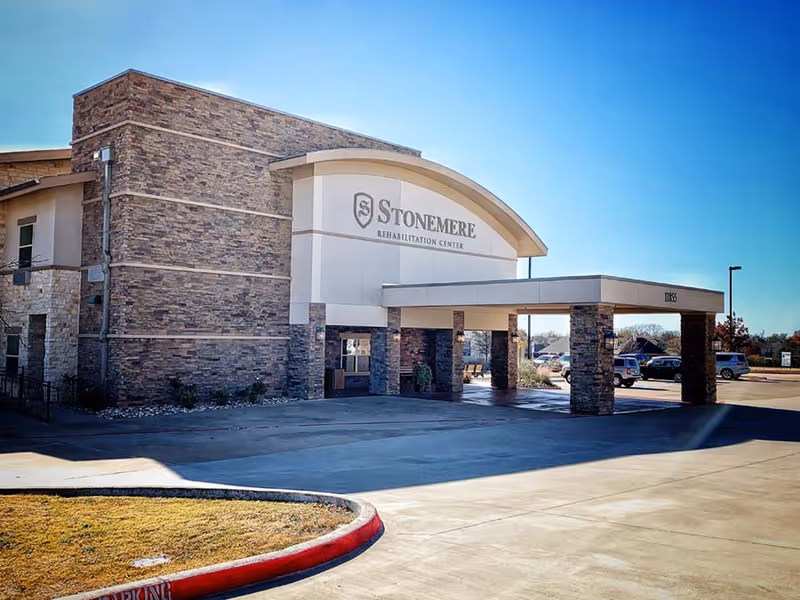 Exterior view of Stonemere Rehabilitation Center building with stone and beige facade, covered entrance supported by stone pillars, and a clear blue sky.