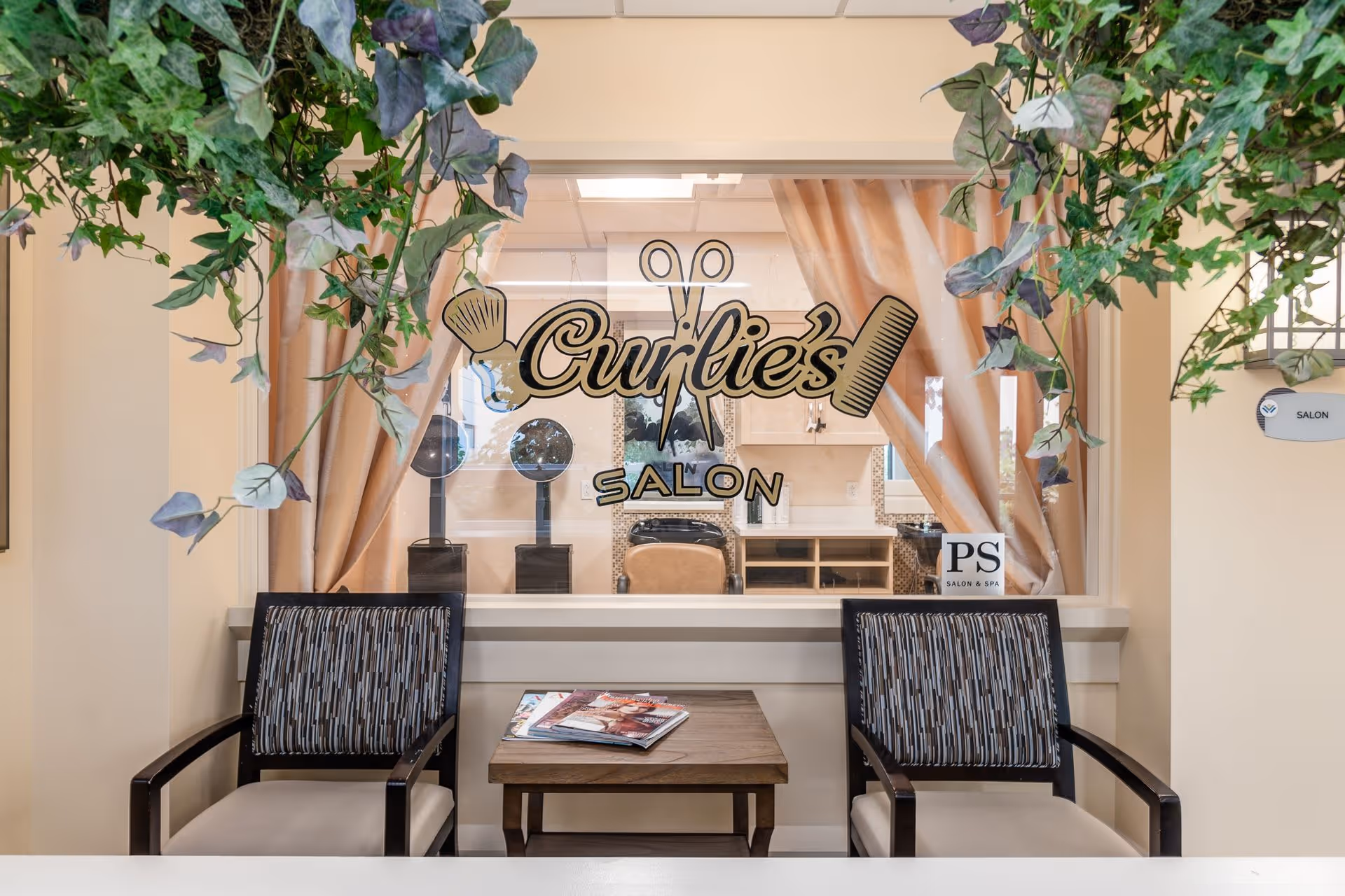 Waiting area of Curlie's Salon with two patterned chairs and a wooden table holding magazines, viewed through a glass window with the salon's name and icons of scissors, a brush, and a comb. Green plants hang from above, and the salon interior with hairdryers and a chair is visible behind the glass.