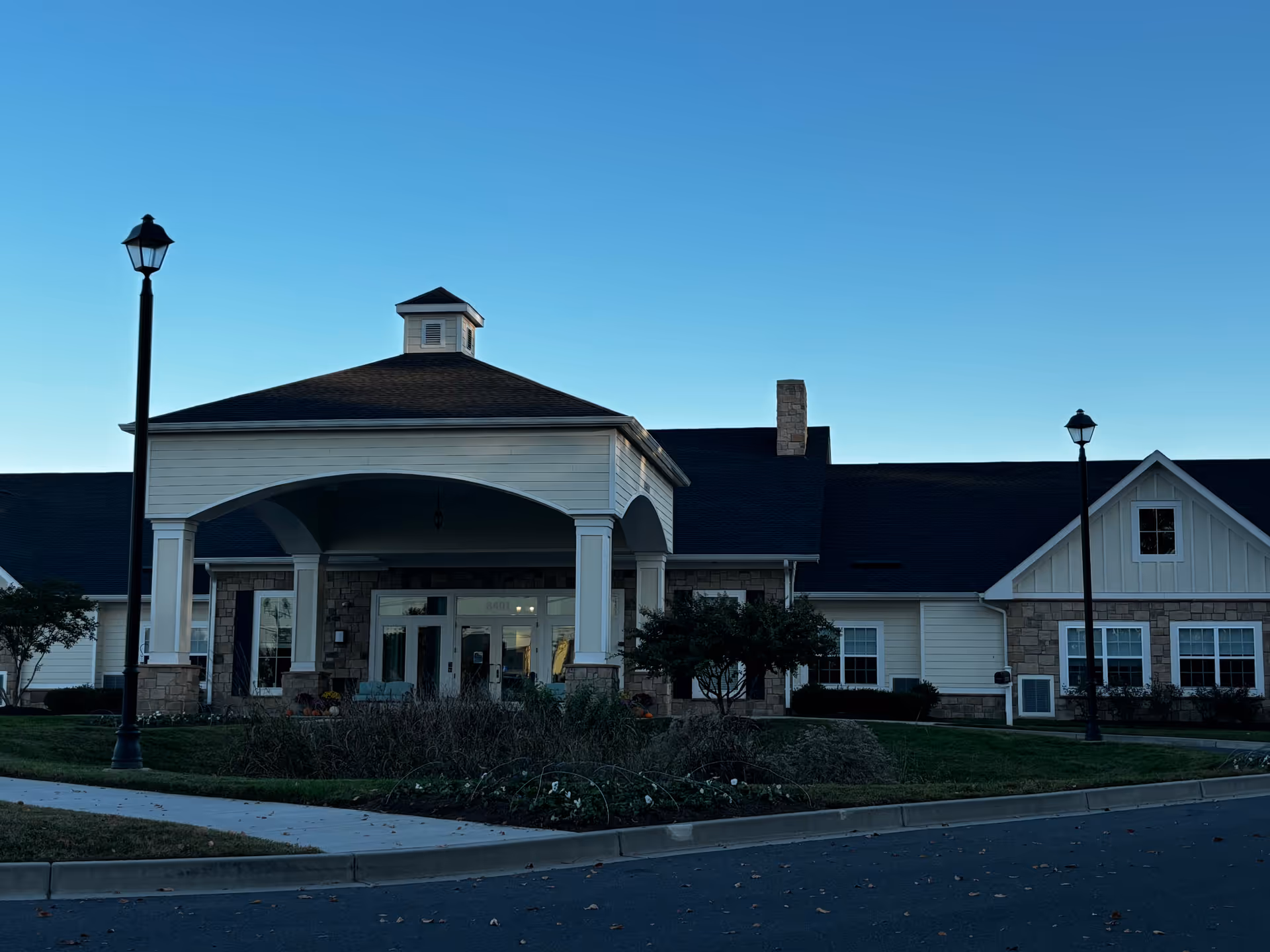 Front entrance of a memory care building with a covered porte-cochère, landscaping, and two lampposts under a clear sky.