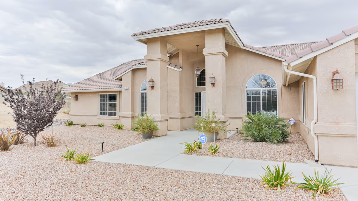 Exterior view of a single-story beige stucco building with a tiled roof, featuring a covered entrance supported by columns. The surrounding landscape is covered with gravel and sparse desert plants, under a cloudy sky.