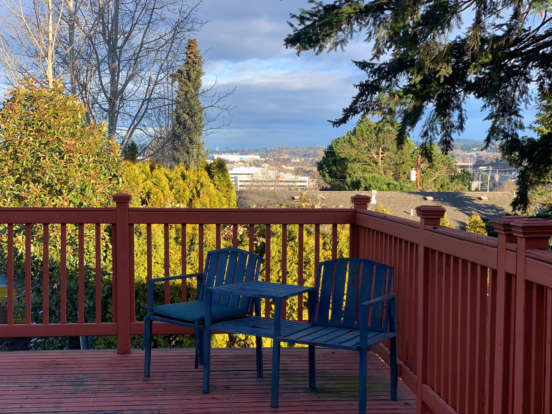 Outdoor wooden deck with red railing, two dark blue chairs with a small table between them, surrounded by green trees and bushes with a view of rooftops and distant cityscape under a partly cloudy sky.