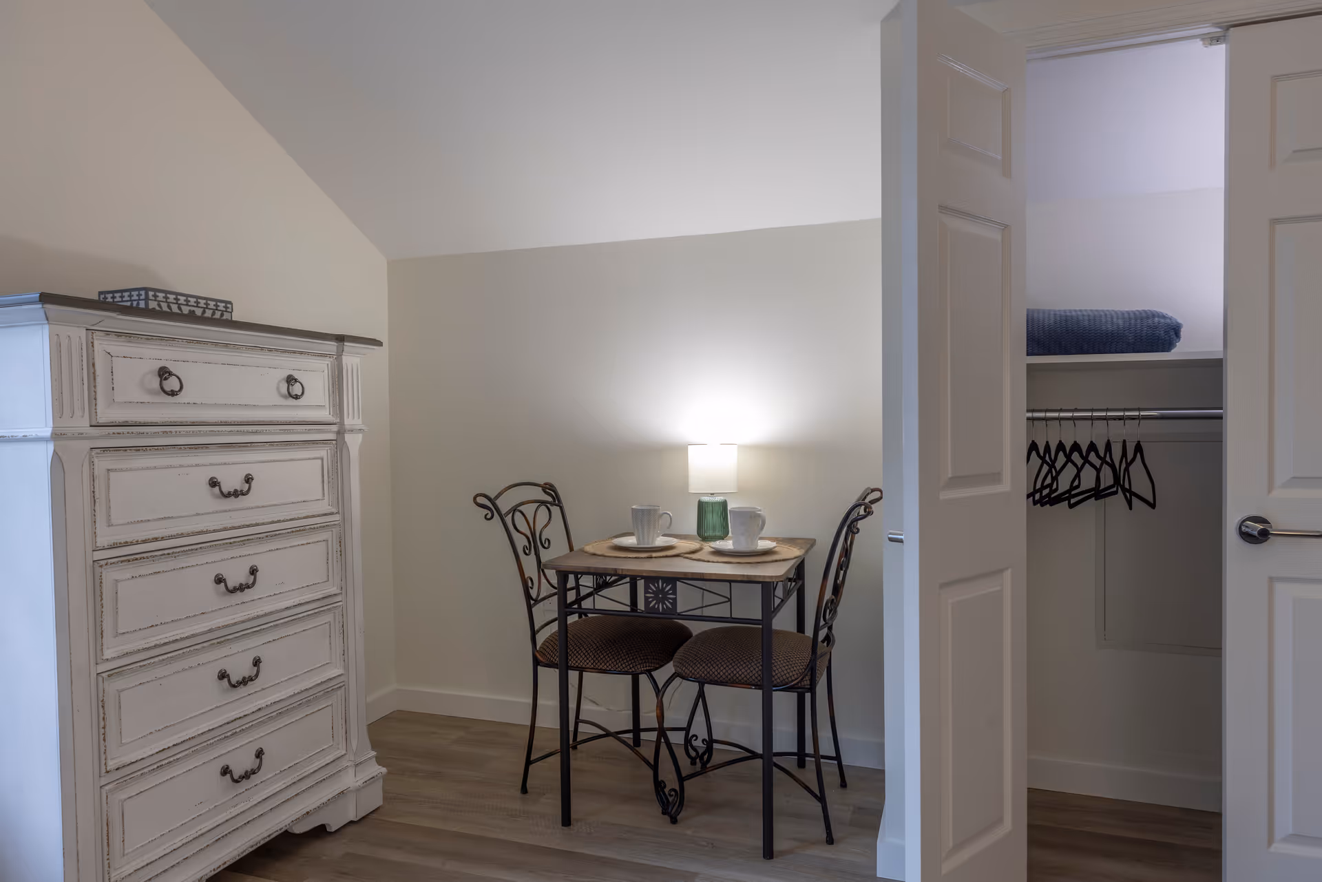 A small dining area with a square table set for two with cups and placemats, two wrought iron chairs with cushioned seats, a white distressed chest of drawers on the left, and an open closet with hangers and a folded blanket on the shelf inside.