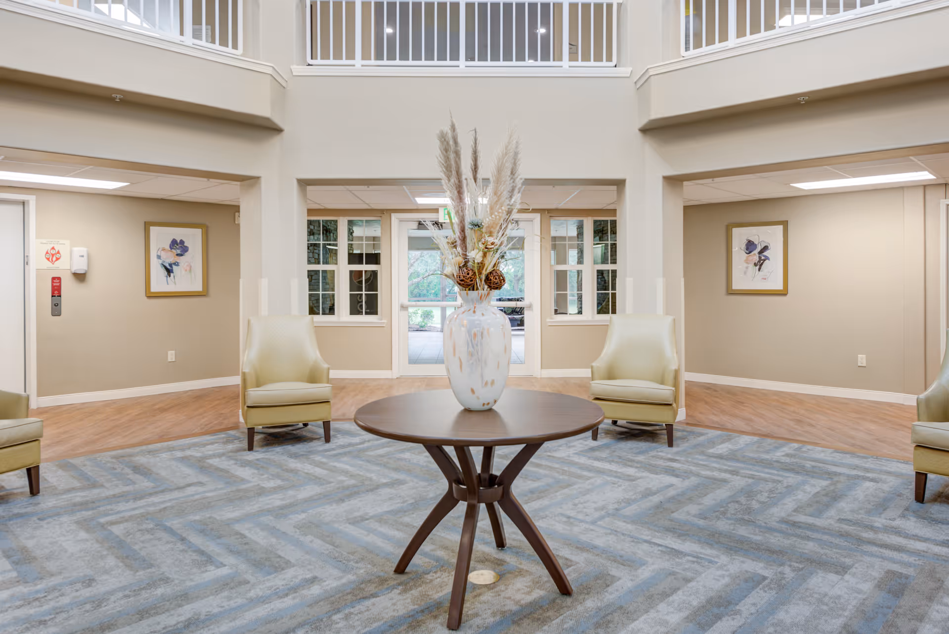 A spacious and well-lit common area with a round wooden table in the center holding a large decorative vase filled with dried plants. Surrounding the table are four light green armchairs placed on a blue patterned carpet. The walls are beige with framed floral artwork, and there are large windows and glass doors leading to an outdoor area. The ceiling is high with a white railing balcony above.