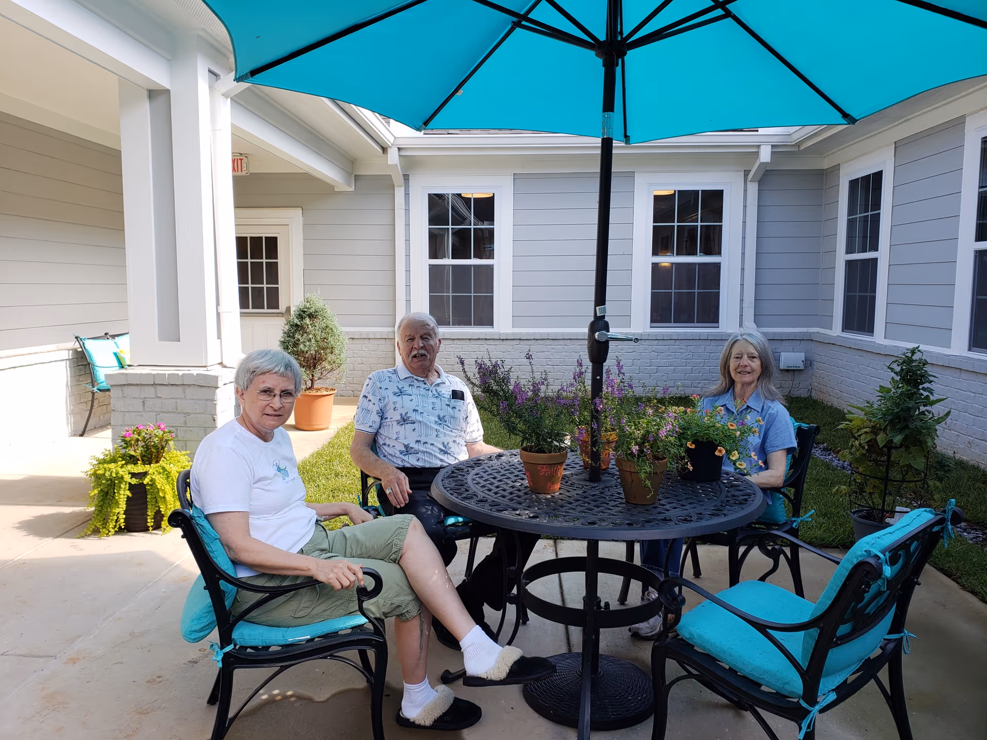 Three elderly people sitting around a round metal table with potted plants on it under a large blue patio umbrella in an outdoor courtyard area with gray siding and white trim on the building walls.