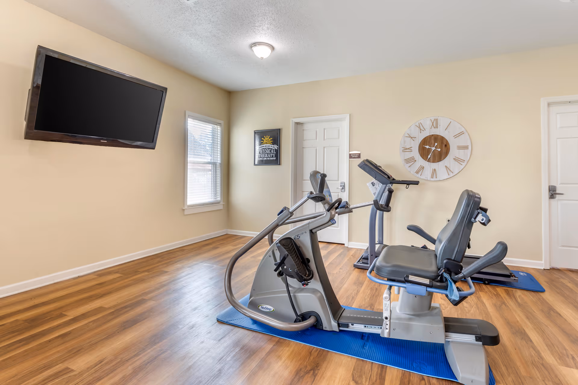 A small exercise room with wooden flooring, beige walls, and a ceiling light. The room contains two pieces of exercise equipment on blue mats, a recumbent exercise bike and a treadmill. A large flat-screen TV is mounted on the wall to the left, and there is a window with blinds next to it. On the back wall, there is a large decorative clock and a framed sign that reads 'Physical Therapy Adds Life to Days'. Two white doors are also visible on the back wall.