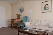 A cozy living room area with a white patterned sofa, a wooden coffee table holding a tissue box and some books, a turquoise armchair, and a small wooden table with two wooden chairs. The walls are decorated with a floral plate, a framed portrait, and a small sign.