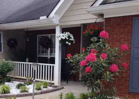 Front porch of a senior living facility with brick exterior, white railing, and hanging flowers.