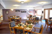 A dining room in a senior living facility with several wooden tables and chairs arranged neatly. Each table is set with folded napkins, plates, and cups. The room has soft purple walls, wall decorations, and large windows allowing natural light to enter.