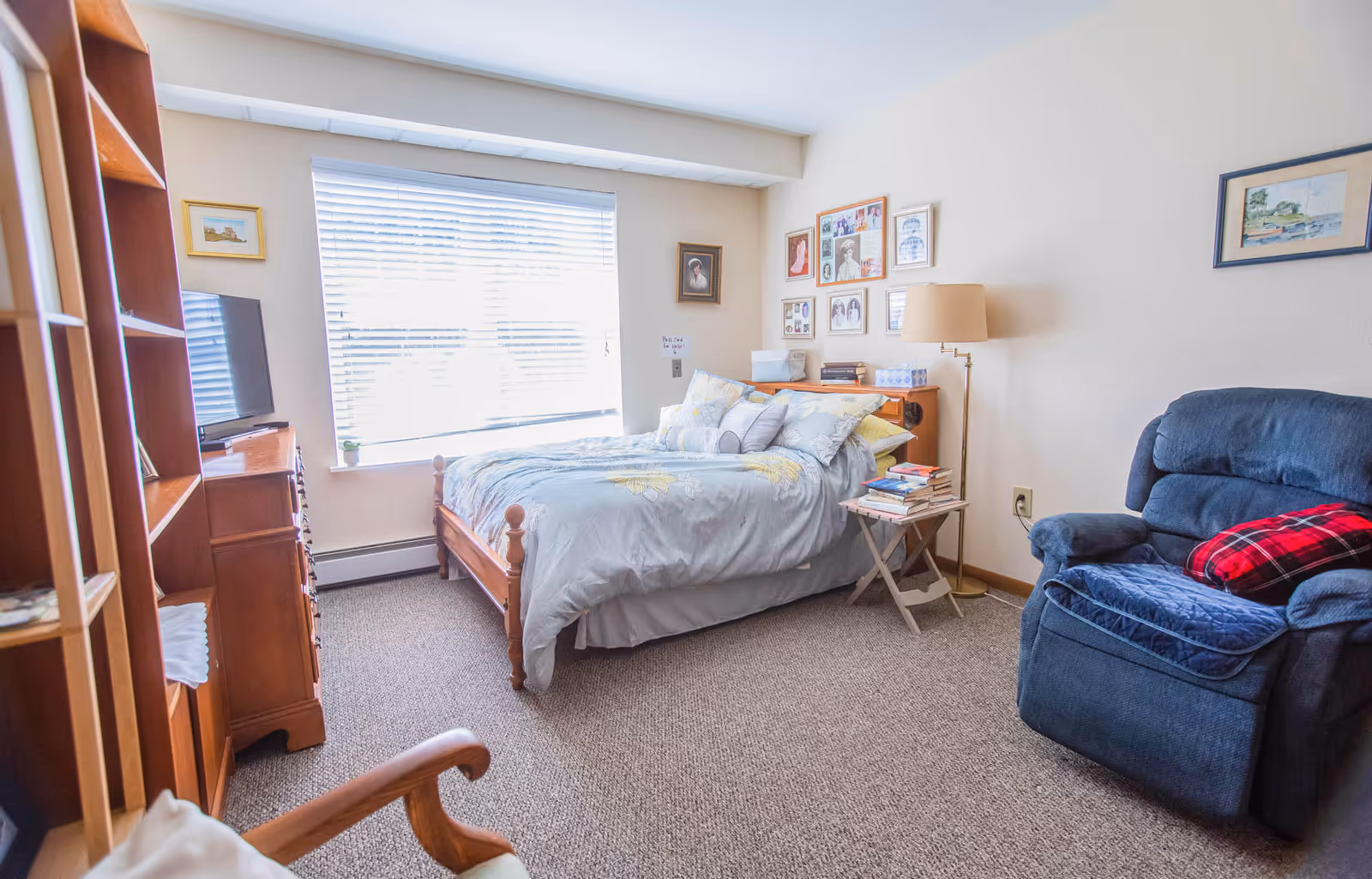A cozy bedroom with a wooden bed covered in light blue bedding with floral patterns, a blue recliner chair with a red plaid pillow, a wooden dresser with a TV on top, a small side table with books, and framed pictures on the walls. A large window with blinds lets in natural light.