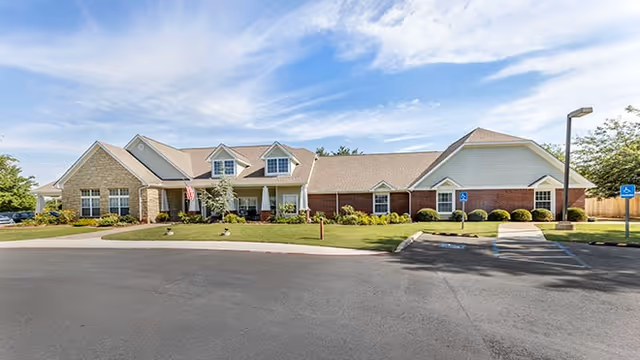 Exterior view of a single-story senior living facility building with a combination of brick and siding, multiple windows, and a front entrance with a covered porch. The building is surrounded by a well-maintained lawn, shrubs, and a paved driveway with handicap parking spaces. The sky is partly cloudy.
