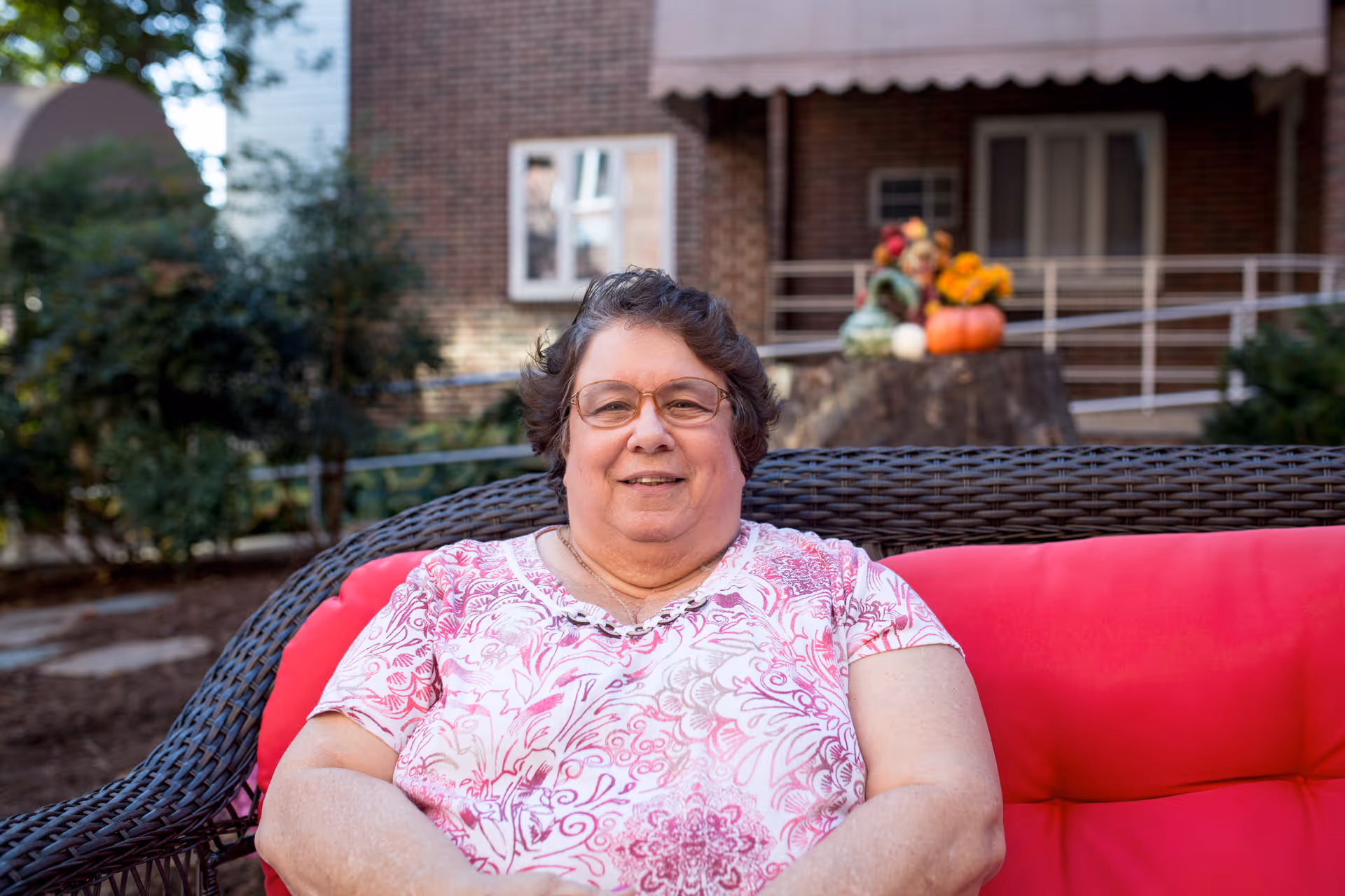 A smiling elderly woman wearing glasses and a pink patterned shirt is sitting on a red cushioned outdoor wicker sofa. Behind her, there is a brick building with a window and a porch railing, along with some autumn-themed decorations including pumpkins and flowers.