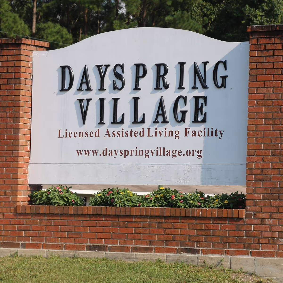 A large outdoor sign for Dayspring Village, a licensed assisted living facility, mounted on a brick structure with flowers planted at the base and trees in the background.