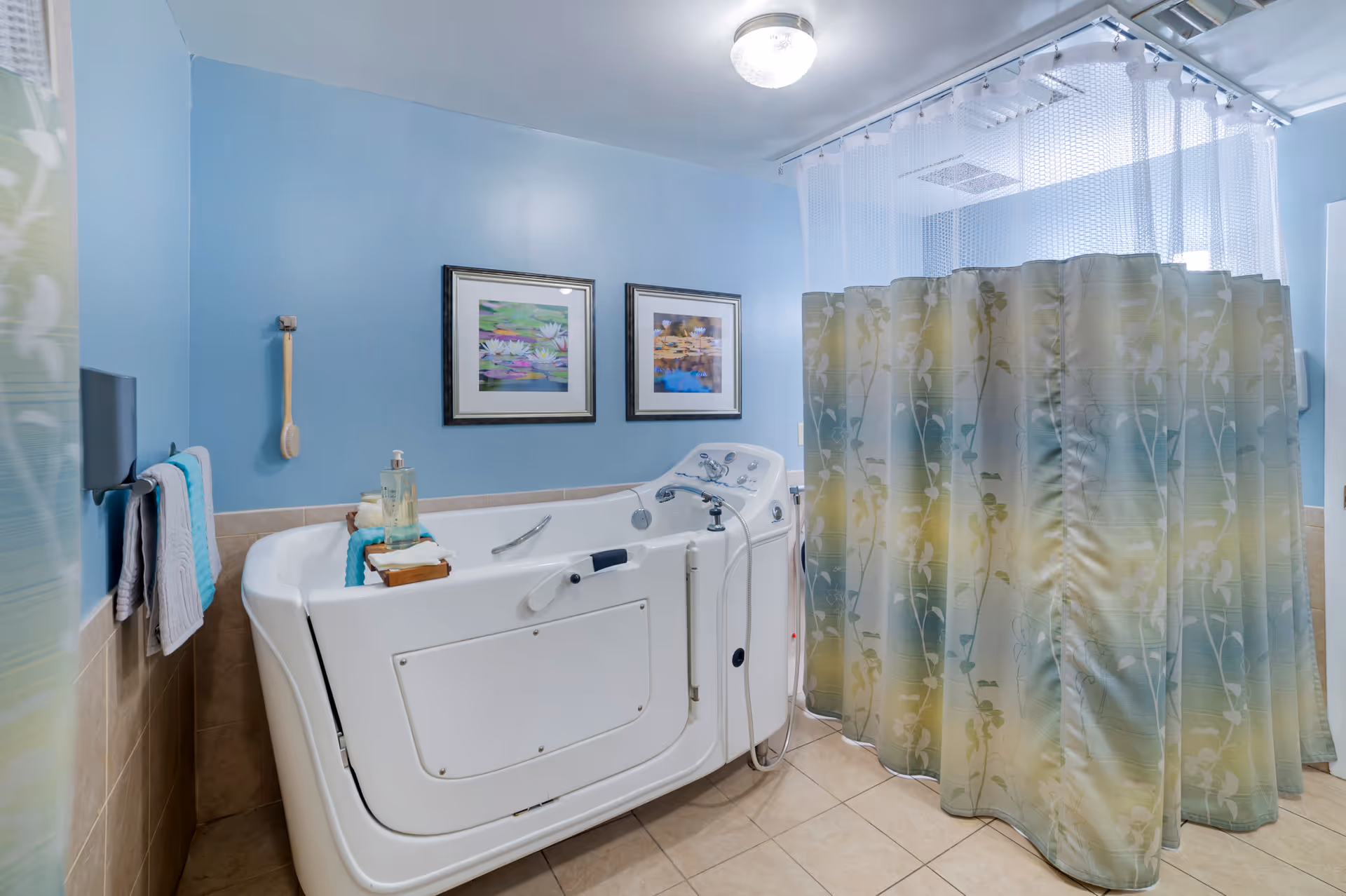 A bathroom with light blue walls and beige tiled floor featuring a white walk-in bathtub with a door, grab bar, and handheld showerhead. Two framed pictures of water lilies hang on the wall above the tub. A towel rack with folded towels and a bath brush hang on the left wall. A floral-patterned shower curtain surrounds the bathtub area.