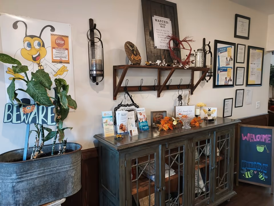 Interior corner of a senior living facility with a decorative cabinet topped with brochures, autumn-themed decorations, and framed quotes. Above the cabinet is a wooden shelf with hooks holding keys and decorative items. To the left, a large potted plant stands next to a cheerful bee-themed sign that says 'BEWARE'. On the right, a chalkboard sign welcomes visitors to BeeHive Homes of Amarillo.
