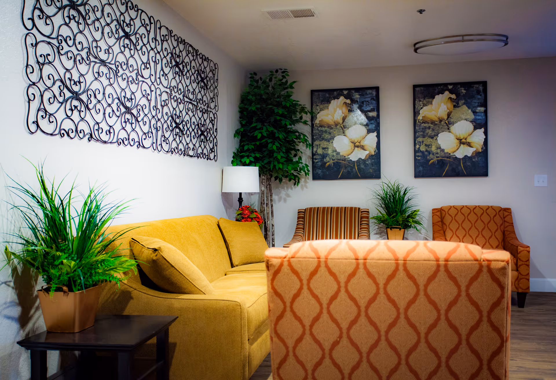 A cozy living room area featuring a mustard yellow sofa, two patterned armchairs, and a small dark wooden side table with a potted green plant. The walls are decorated with two floral paintings and an intricate black metal wall art piece. A floor lamp and a tall artificial tree add to the room's decor.