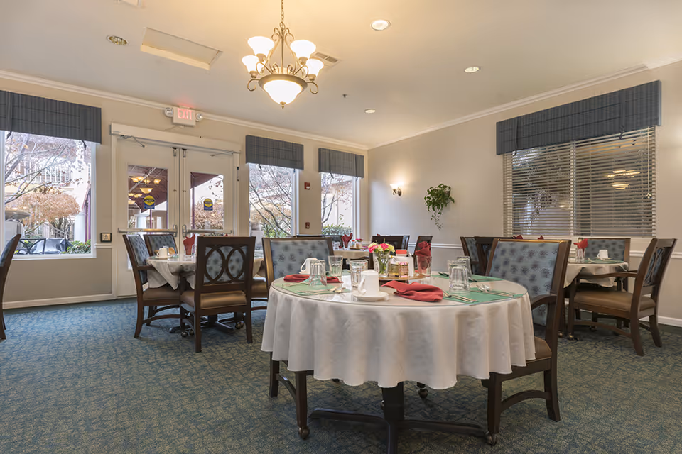 A dining room in a senior living facility with round and square tables covered with white tablecloths, set with cups, glasses, silverware, and red napkins. The room has large windows with blue valances, a chandelier, and a blue carpeted floor. Outside trees with autumn leaves are visible through the windows.