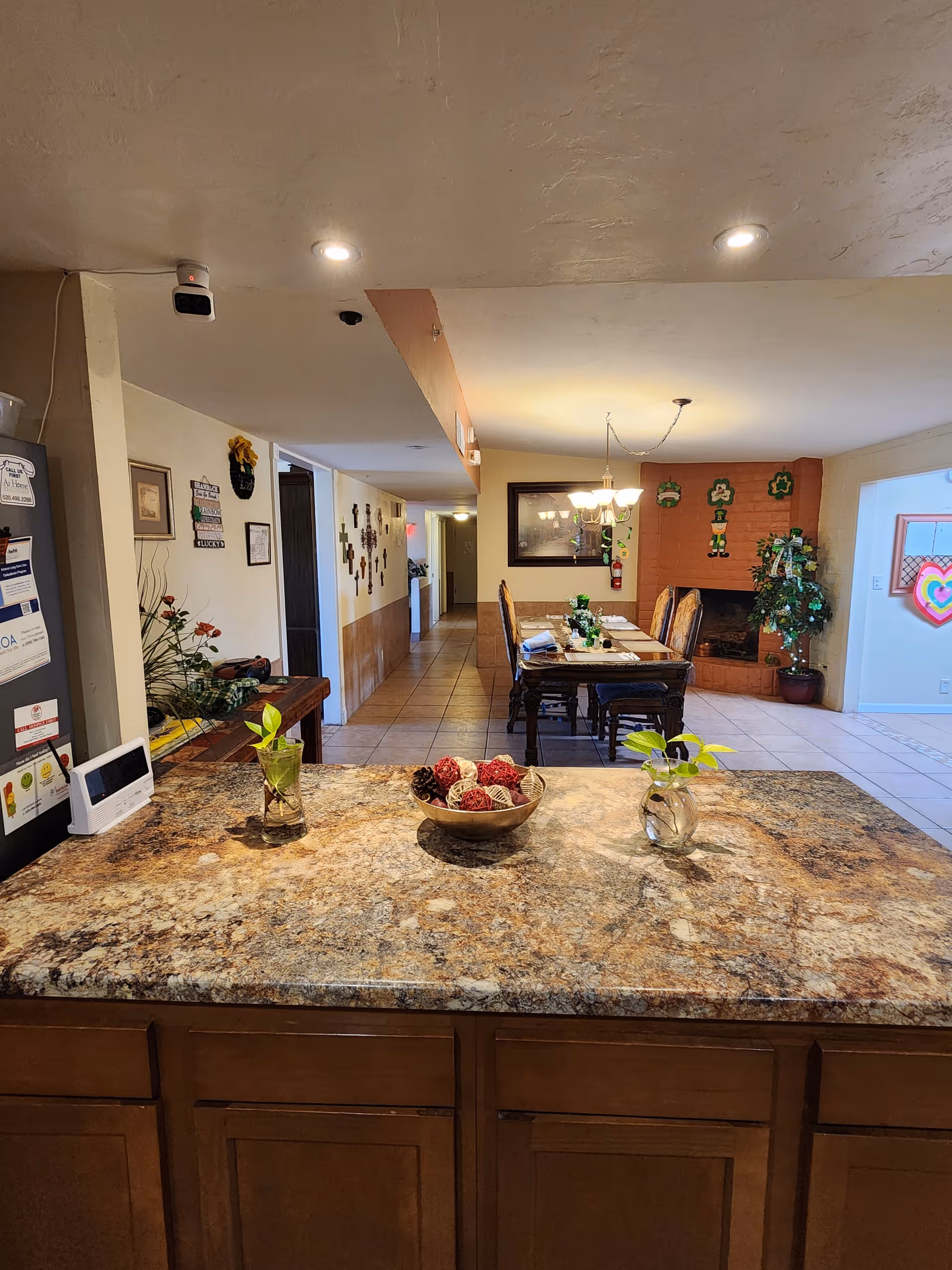 View from a kitchen counter with a granite countertop featuring two small glass vases with green plants and a bowl with decorative red and brown balls. In the background, there is a dining area with a wooden table and chairs, a chandelier, a brick fireplace decorated with St. Patrick's Day decorations, and a hallway leading to other rooms.