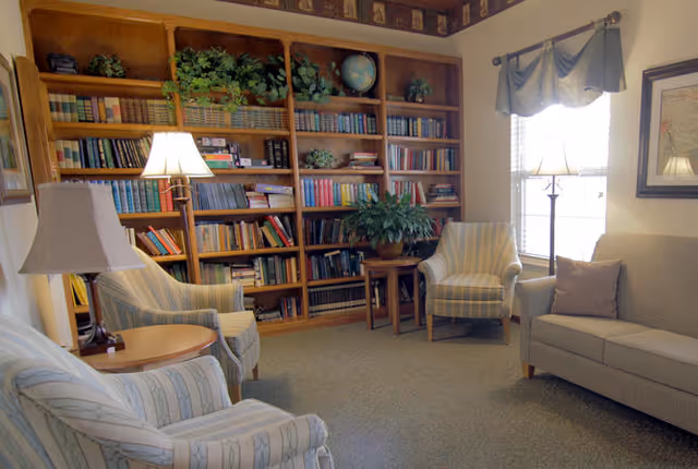 A cozy living room with a large wooden bookshelf filled with books and plants. The room has three upholstered armchairs and a sofa, all arranged around two side tables with lamps. A window with curtains and a framed picture are visible on the wall.
