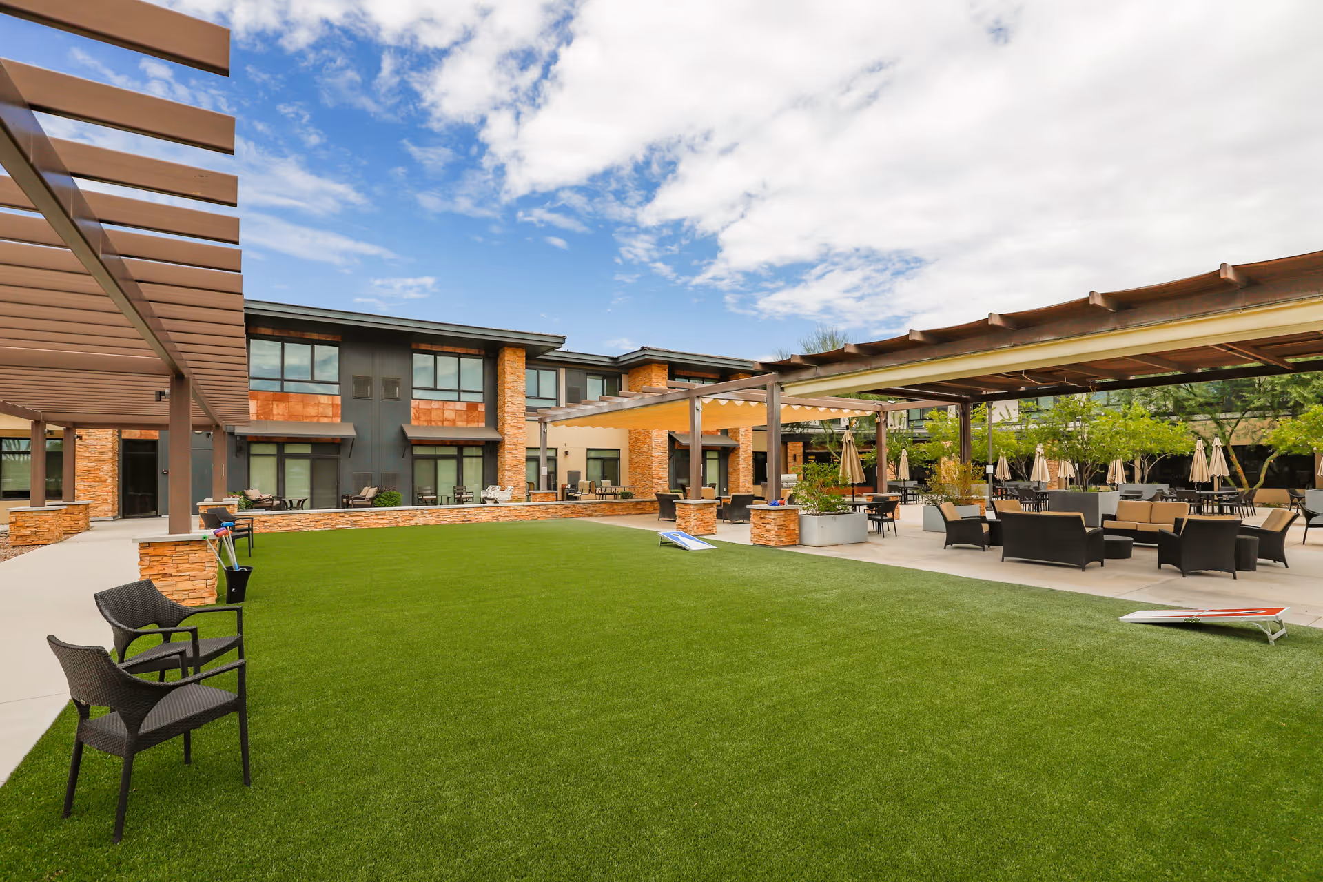 Outdoor courtyard area at Merrill Gardens at Anthem featuring a large green lawn, several seating areas with chairs and tables under pergolas, and a two-story building in the background under a partly cloudy sky.