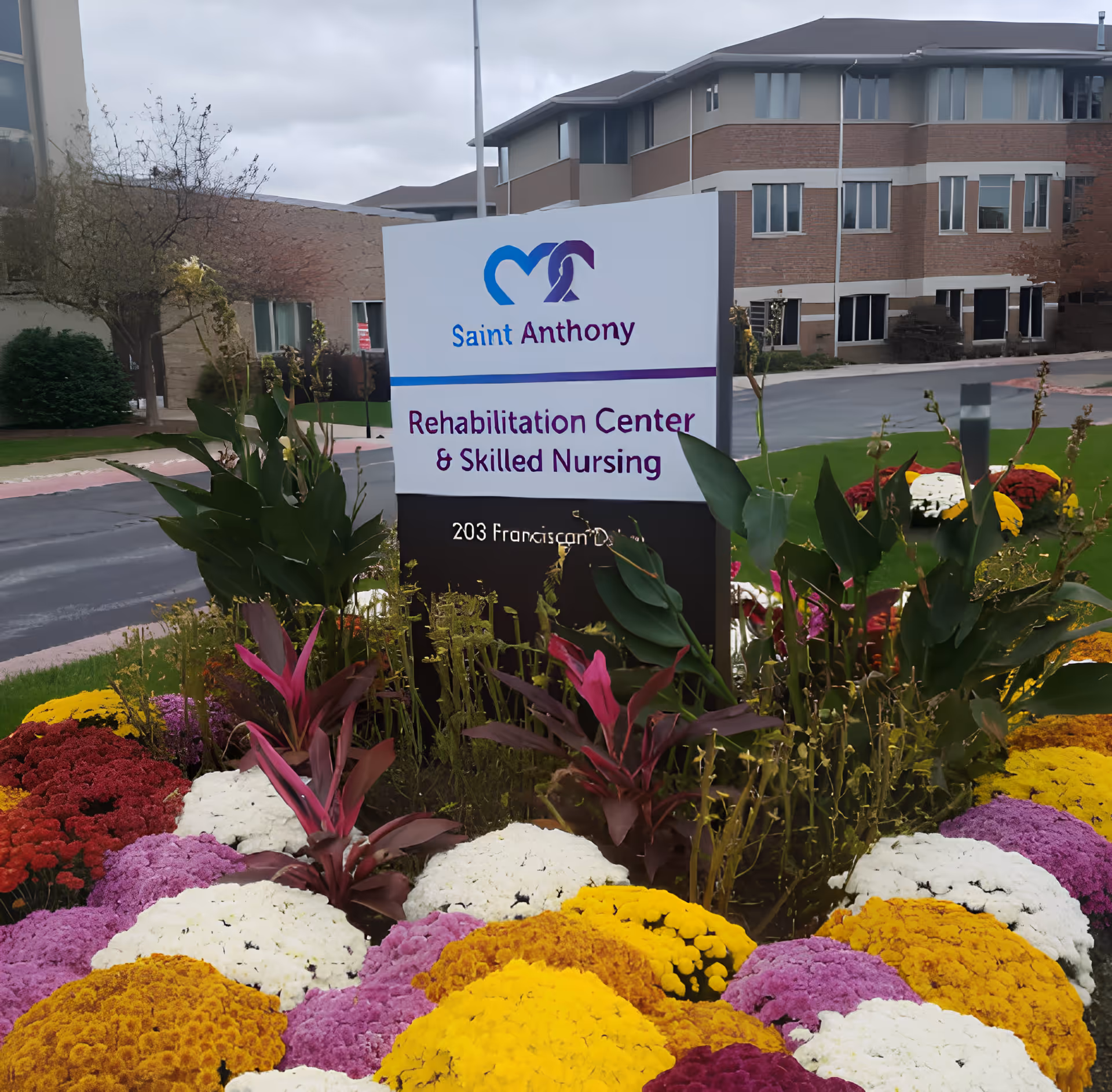 Outdoor view of Saint Anthony Rehabilitation Center & Skilled Nursing sign surrounded by colorful flowers with a multi-story brick building in the background under a cloudy sky.