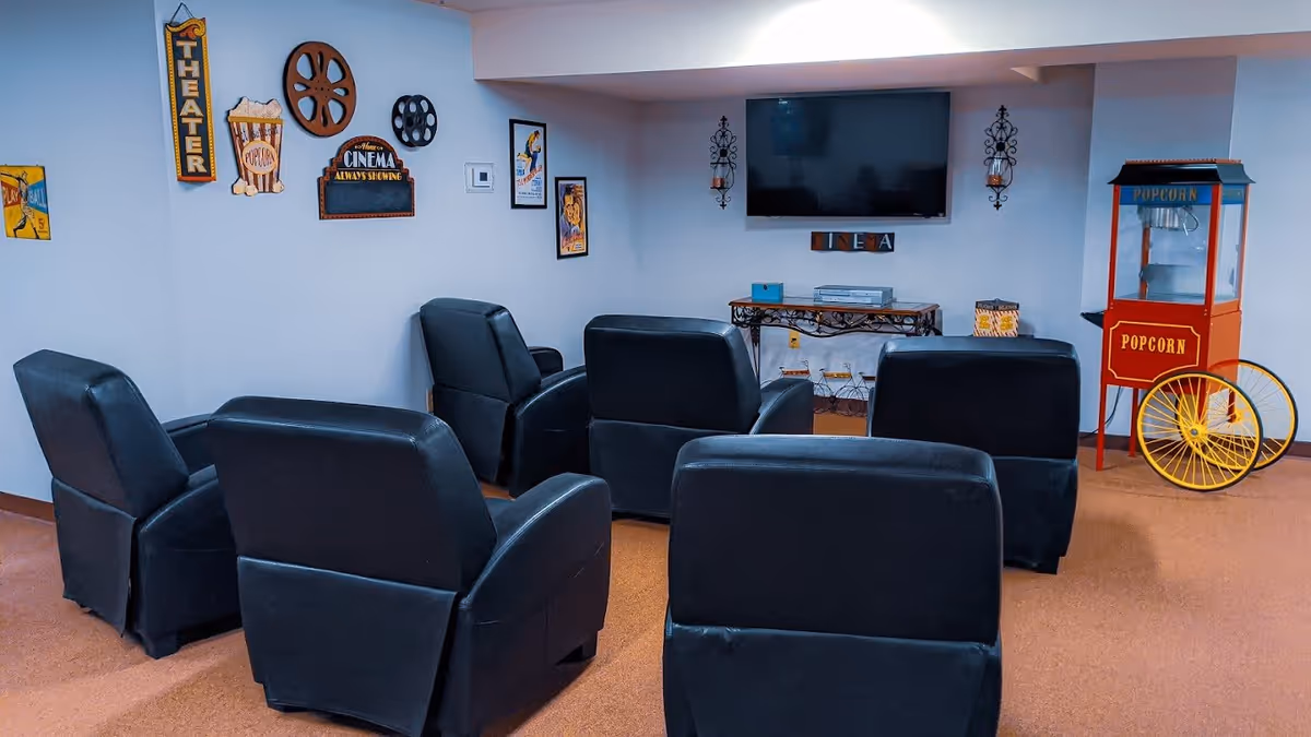 A cozy movie room with six black recliner chairs arranged in two rows facing a wall-mounted flat screen TV. The walls are decorated with vintage cinema-themed signs and posters. To the right, there is a red and yellow popcorn machine on wheels.