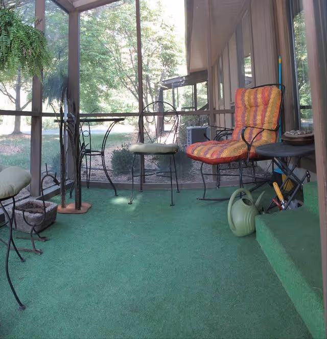 A screened-in porch area with green carpet flooring, featuring a cushioned chair with a colorful orange and red pattern, a small round table, a watering can, and various garden tools. Outside the screen, trees and greenery are visible.
