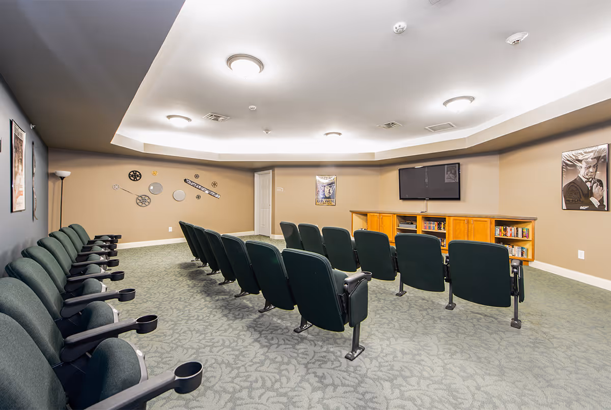 A small theater room with two rows of green cushioned seats equipped with cup holders facing a wall-mounted flat screen TV. The room has beige walls decorated with movie-themed posters and film reel wall art. There is a wooden cabinet beneath the TV holding DVDs and other media items. The carpet has a subtle leaf pattern, and the ceiling features recessed lighting.
