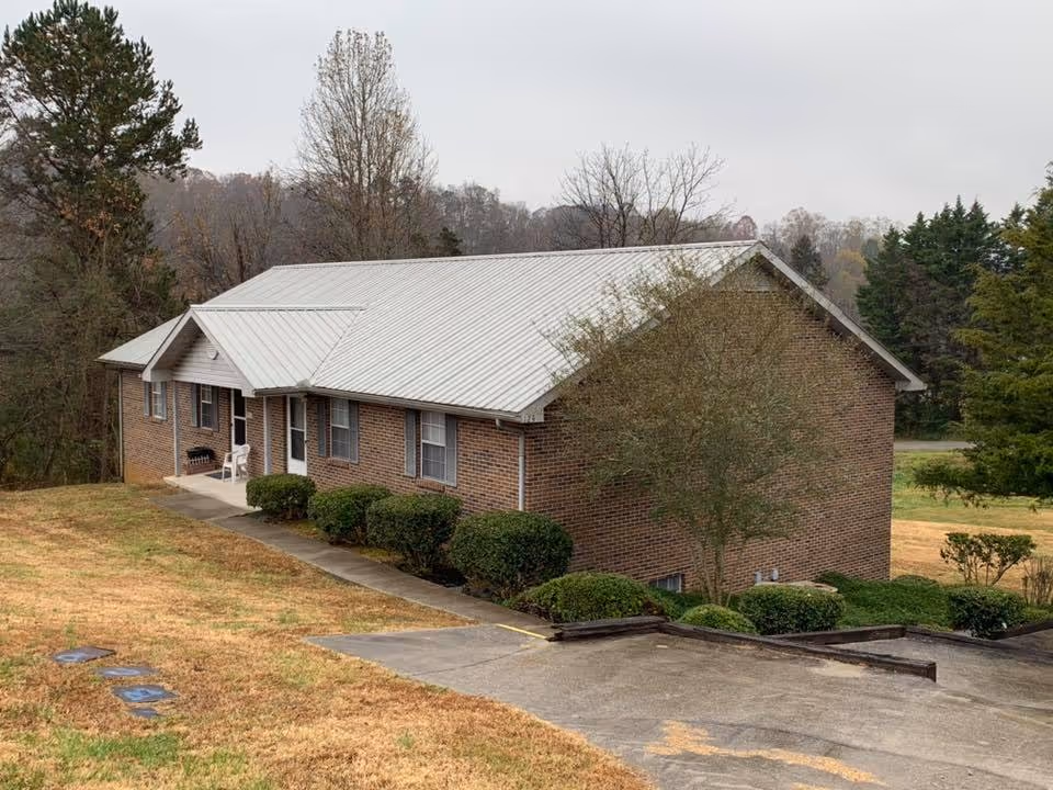 Brick single-story building with a metal roof, front entrance and shrubs on a sloped lawn.