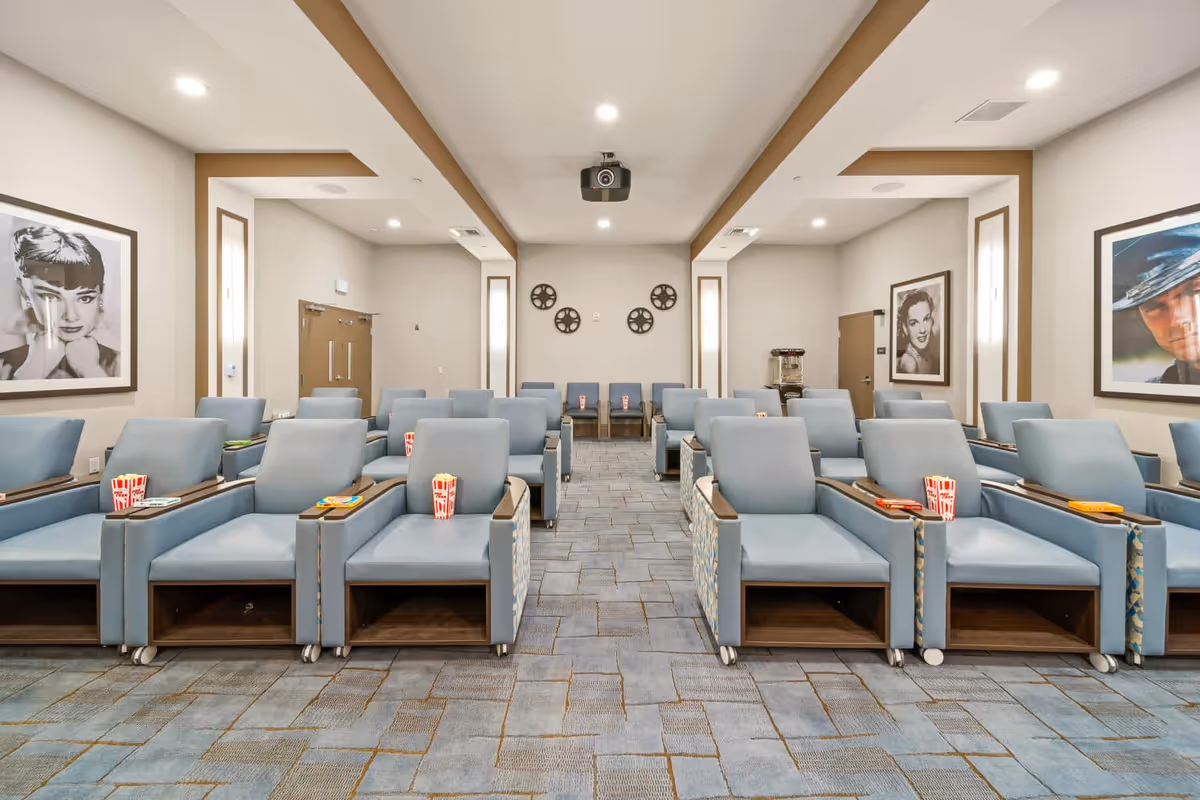 A modern movie theater room with rows of light blue reclining chairs, each with a small table holding popcorn and snacks. The room has a projector mounted on the ceiling, movie reel decorations on the back wall, and black-and-white portraits of classic movie stars on the side walls.