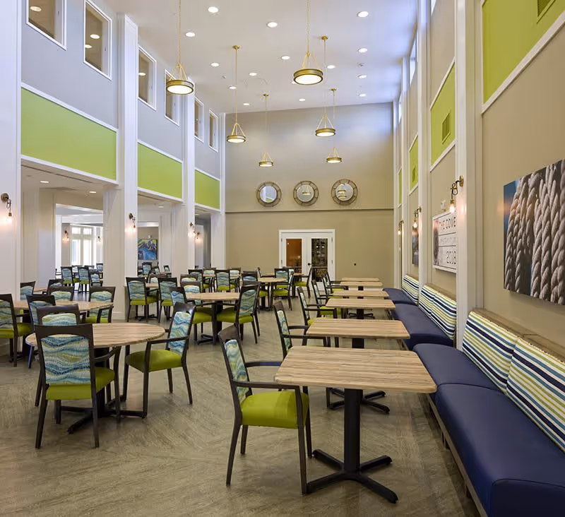 A spacious dining room with multiple wooden tables and green cushioned chairs. The room features high ceilings with hanging pendant lights, large windows on one side, and a long blue cushioned bench with striped pillows along the wall. The walls are painted in neutral tones with green accents and decorative round mirrors above a set of double doors.