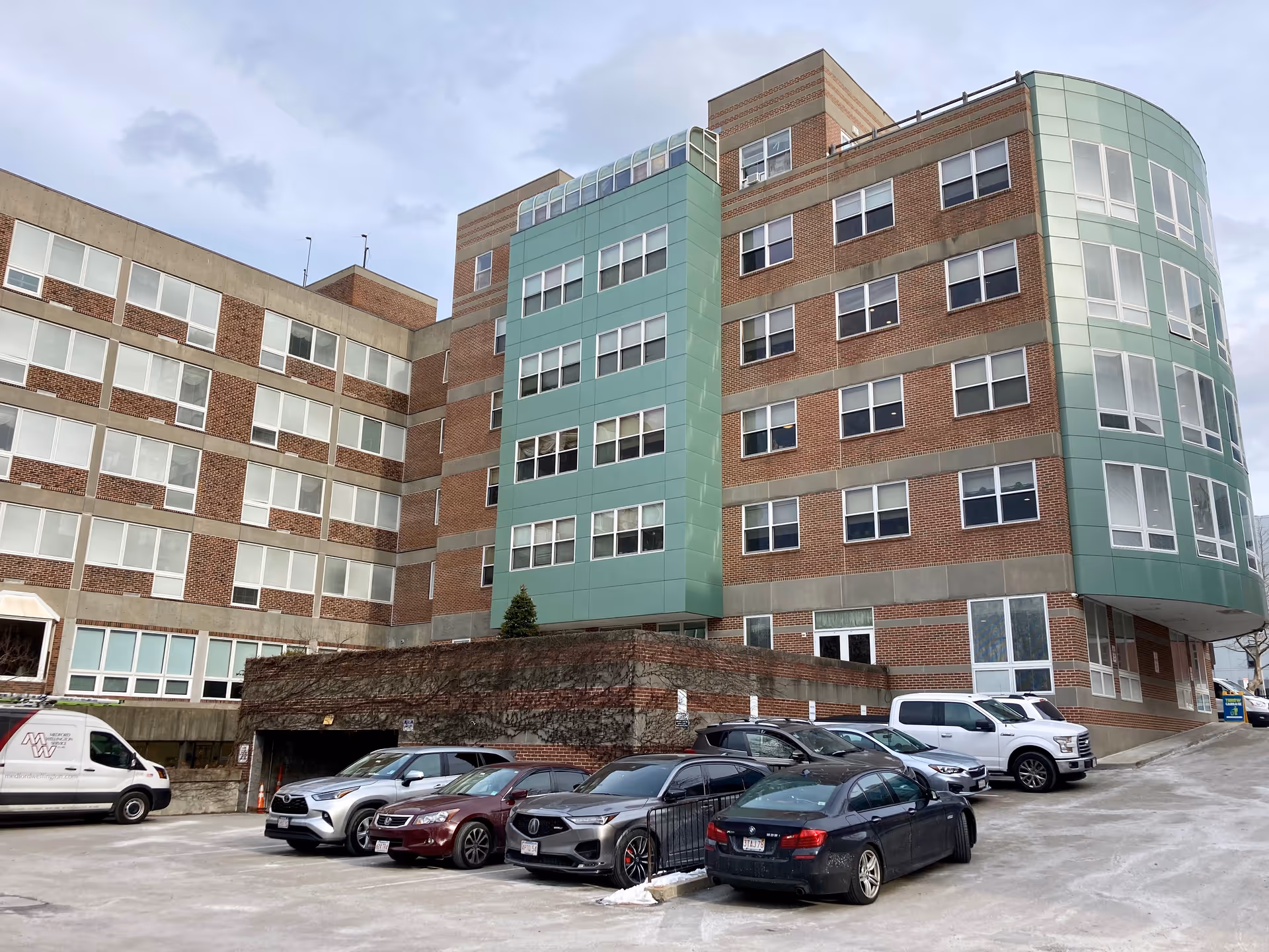 Multi-story brick and green-paneled senior living building with a parking lot and several cars in front under a cloudy sky.