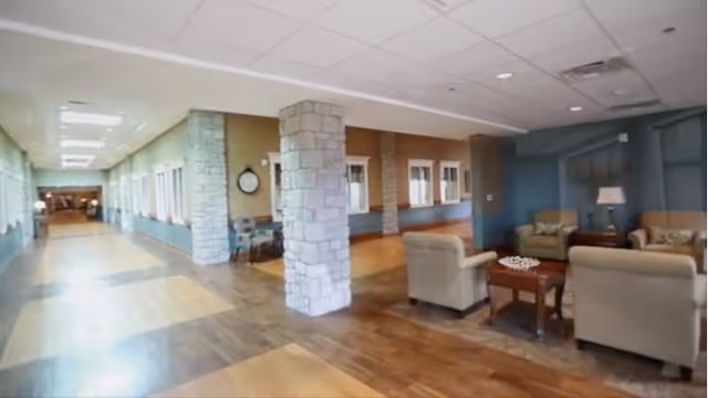 Interior view of a senior living community hallway with stone pillars and a seating area featuring armchairs, a coffee table, and a lamp. The hallway has large windows on one side and a wooden floor with a patterned design.