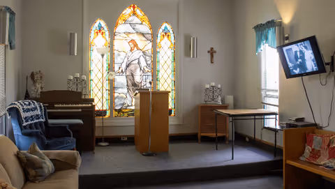 Small interior chapel-like common room with a stained-glass window behind a lectern on a raised platform, piano, seating and a wall-mounted TV.