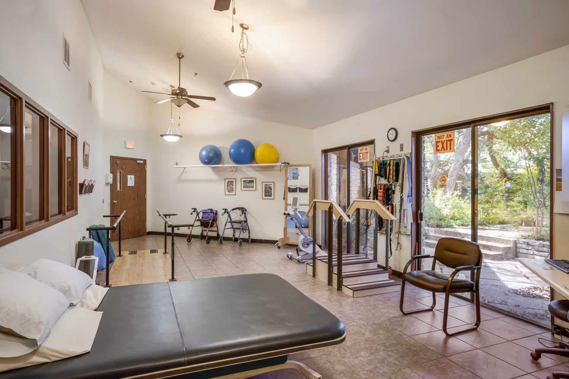 A physical therapy room with exercise equipment including parallel bars, walkers, therapy balls on a shelf, a therapy table with pillows, a chair, and a small set of stairs with handrails. Large windows and glass doors show an outdoor garden area with trees and stone steps. The room has ceiling fans and hanging lights.