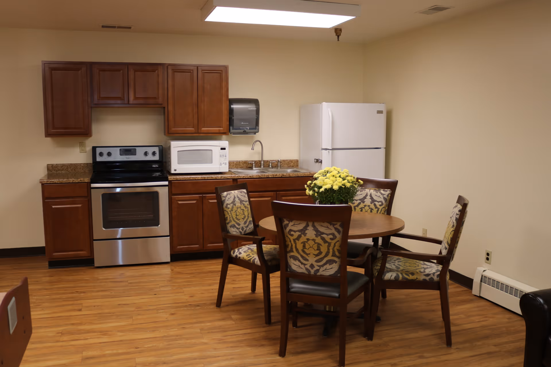 A small communal kitchen with wooden cabinets, a stove, microwave, refrigerator and a round dining table with four chairs and a vase of yellow flowers.