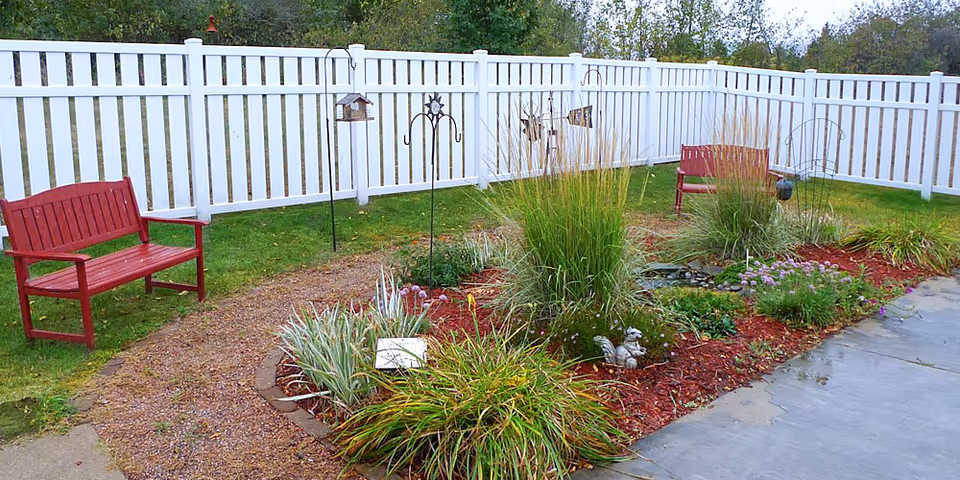 Fenced outdoor garden courtyard with white picket fence, red benches, planted flower beds and a gravel path.