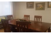 A dining room with a wooden table and six matching wooden chairs. There is a decorative centerpiece on the table. The room has light-colored walls with three framed pictures hanging on the wall. A window with sheer white curtains is visible on the left side.