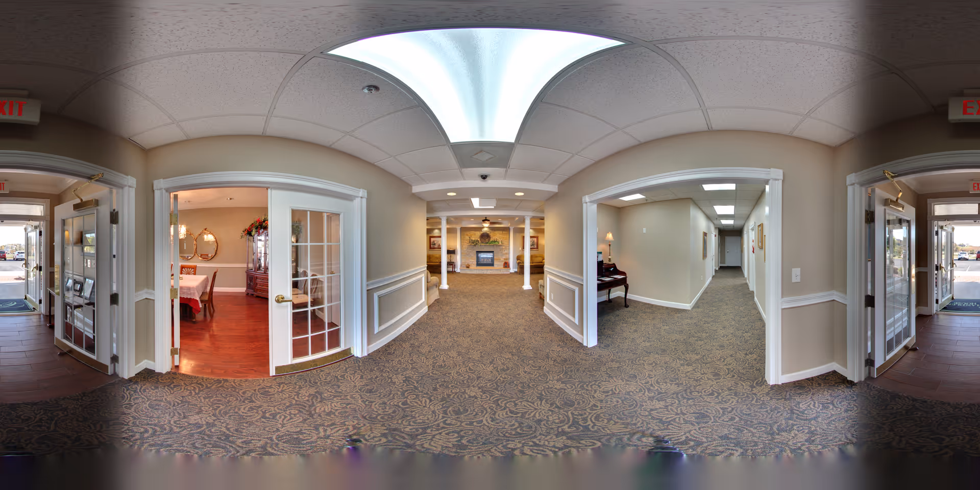 Interior view of a senior living facility hallway with patterned carpet and beige walls. On the left, there is a room with glass double doors leading to a dining area with a table and chairs. Straight ahead is a common area with a fireplace and columns. On the right, there is a hallway with framed pictures and a piano against the wall. Exit signs are visible above the doors at both ends of the hallway.