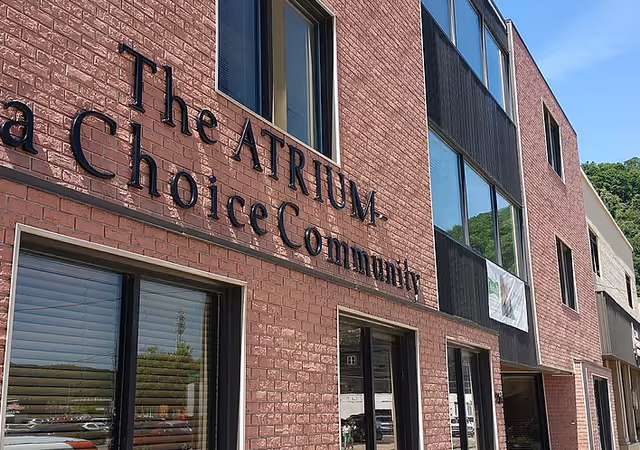 Exterior view of a brick building with large windows and the sign 'The ATRIUM - a Choice Community' mounted on the wall under a clear blue sky.